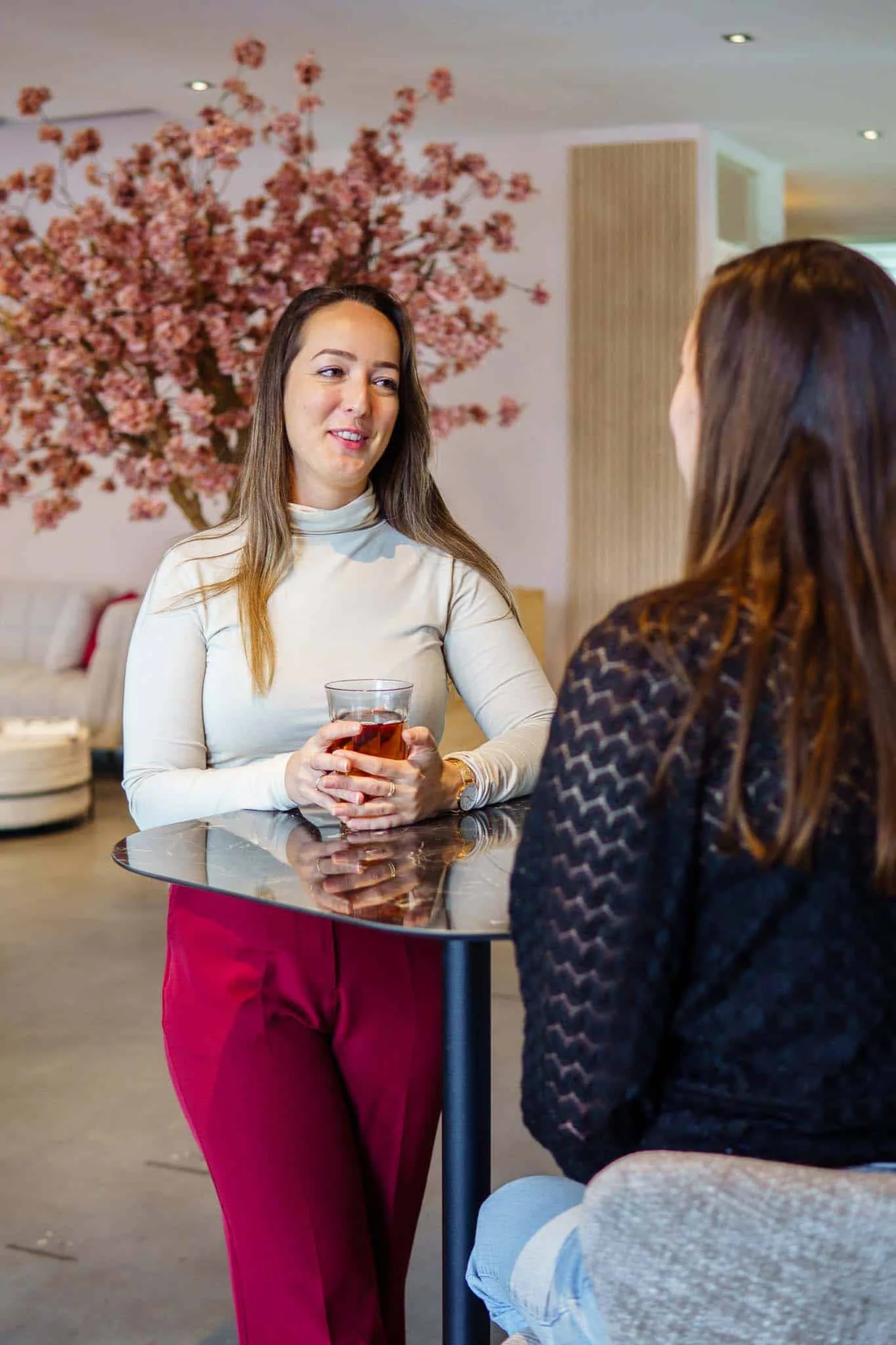 Twee vrouwen in gesprek in een stijlvolle lobby, één met een glas thee, rode broek, en een lachende uitdrukking.