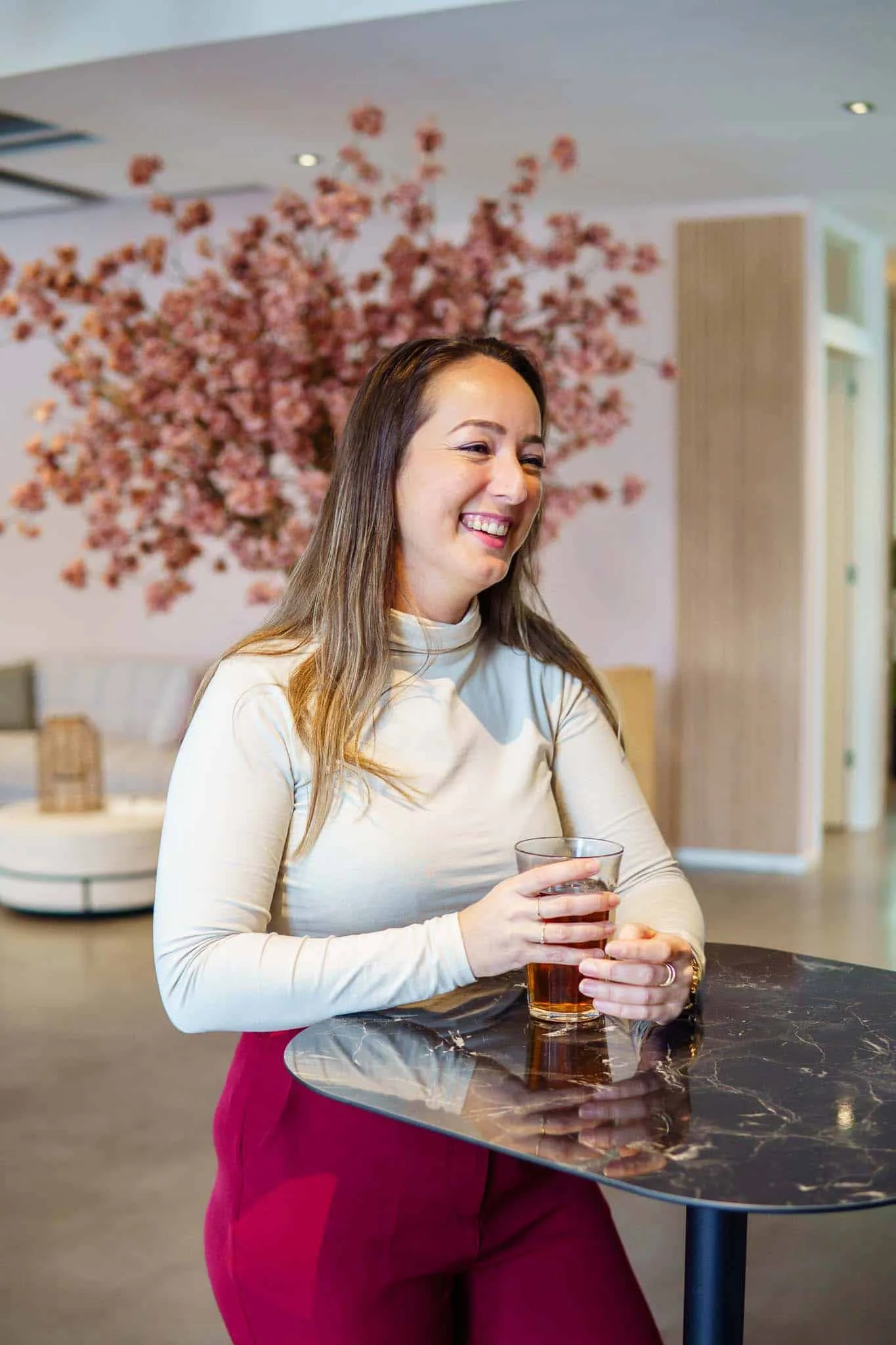 Lachende vrouw geniet van een drankje aan een zwarte tafel in een stijlvolle lobby met bloemdecoratie op de achtergrond. Casual en ontspannen sfeer.