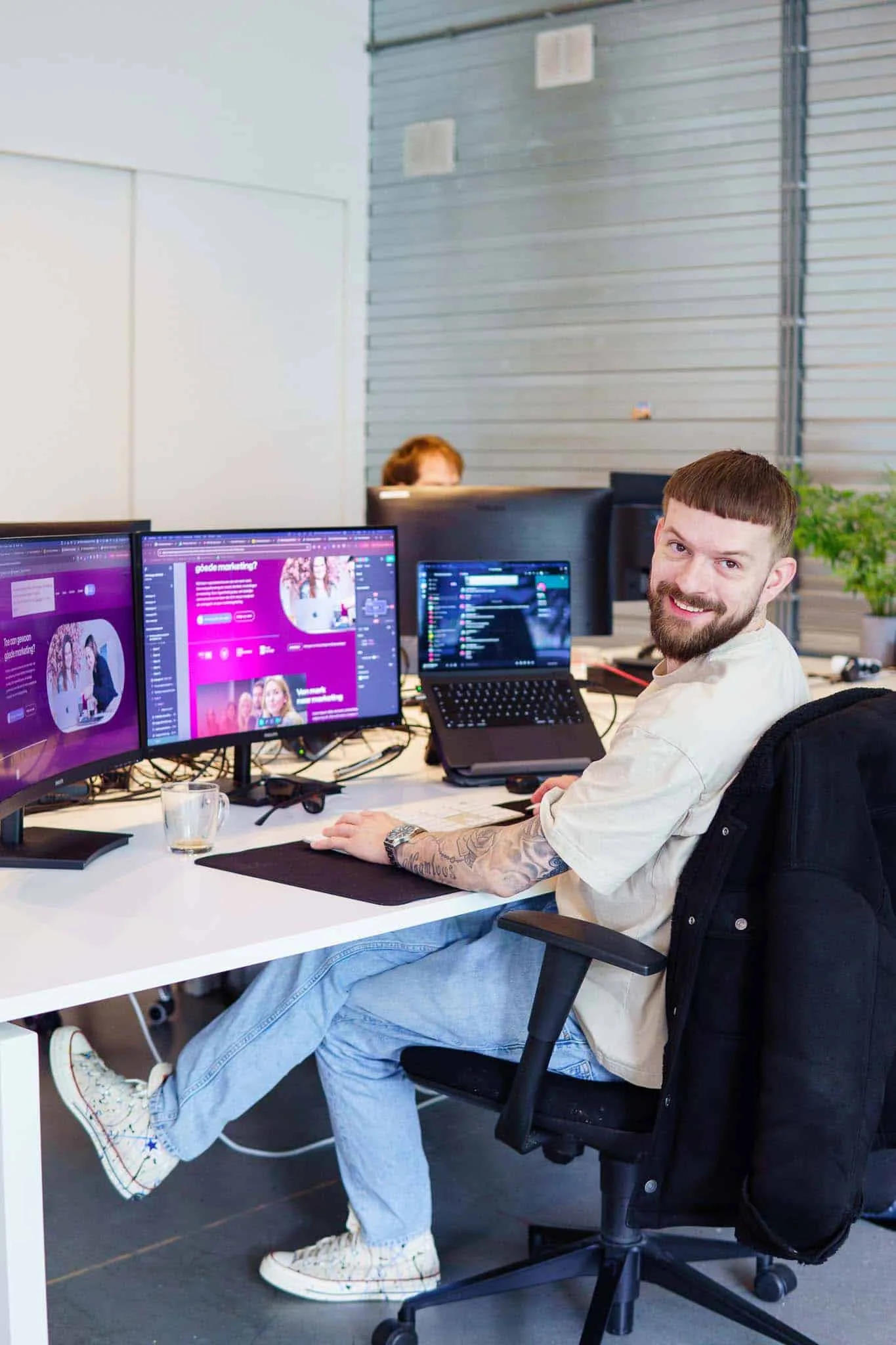 Developer kijkt lachend om vanachter drie schermen met een websiteontwerp
