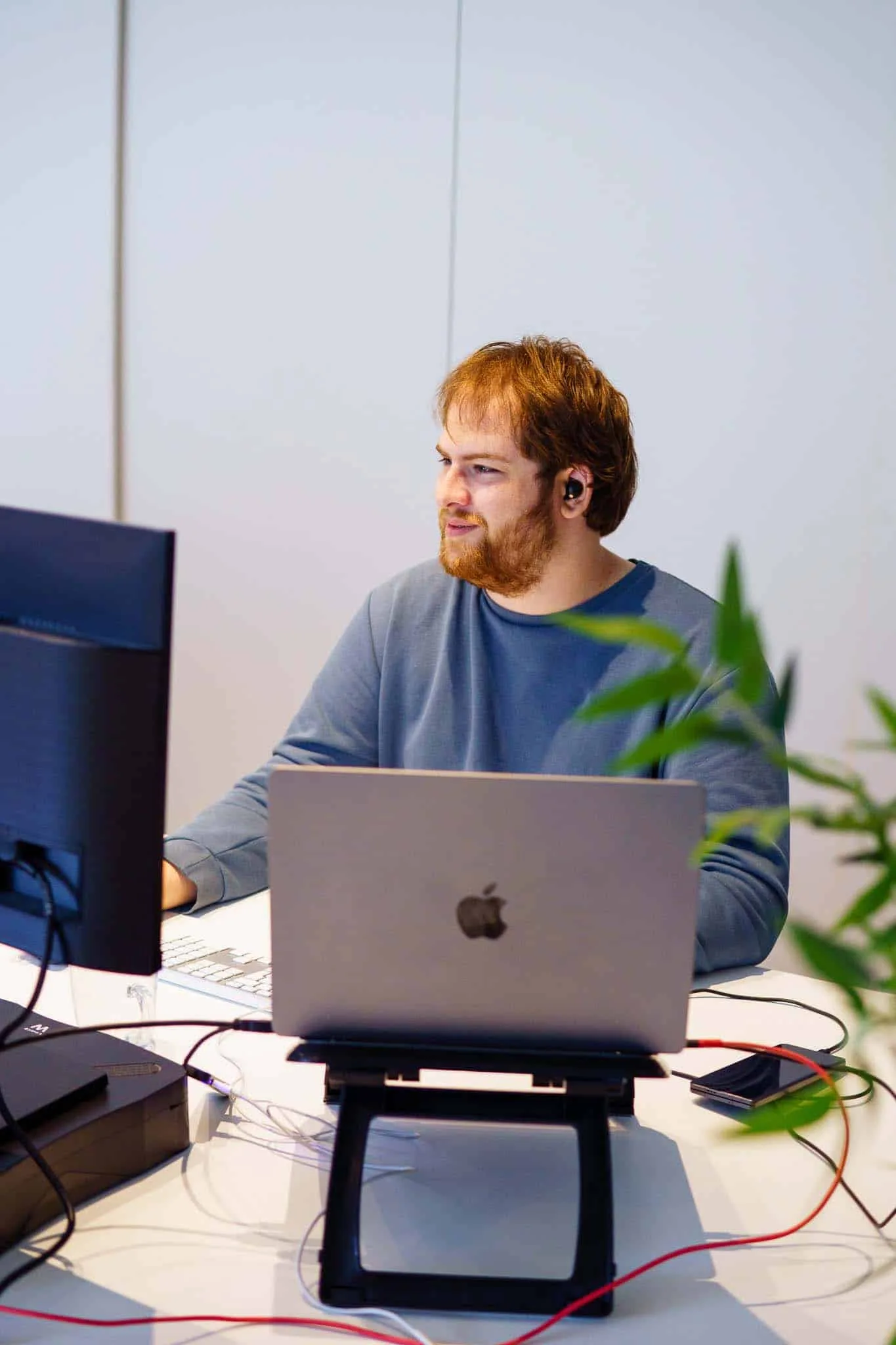 Jonge man met rood haar werkt geconcentreerd achter zijn laptop en monitor op kantoor. Hij draagt oordopjes en heeft een rustige uitstraling.