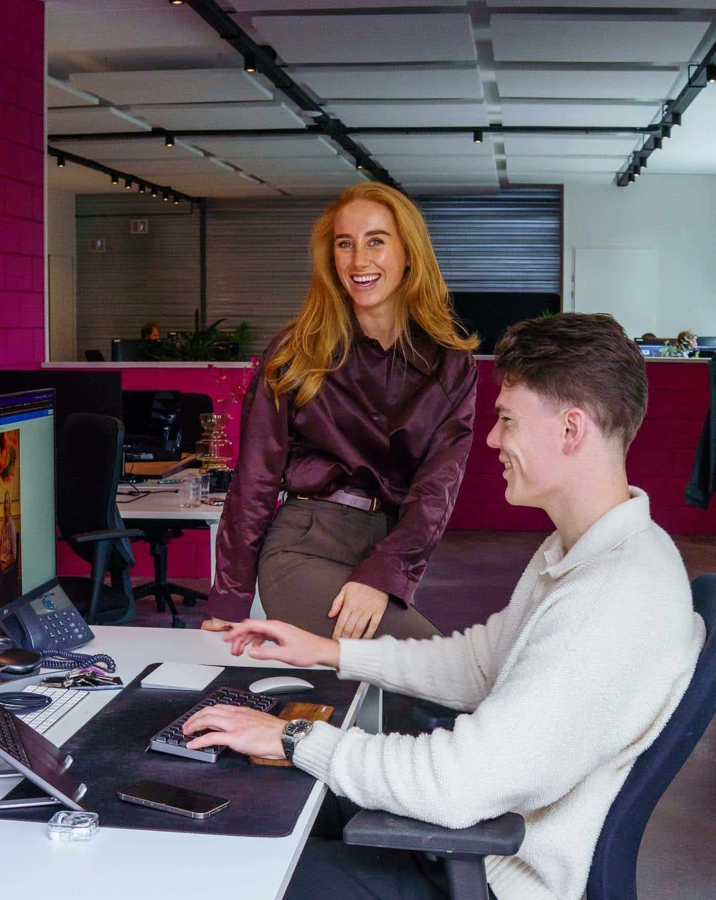 Twee jonge collega's in een modern kantoor: een man typt op een toetsenbord terwijl een vrouw met lang rood haar lachend toekijkt. Samenwerking en teamwerk.