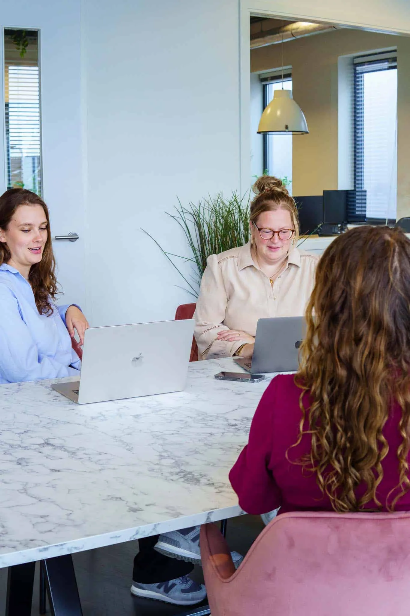 Drie jonge vrouwen werken samen aan een marmeren tafel met laptops in een lichte kantoorruimte.