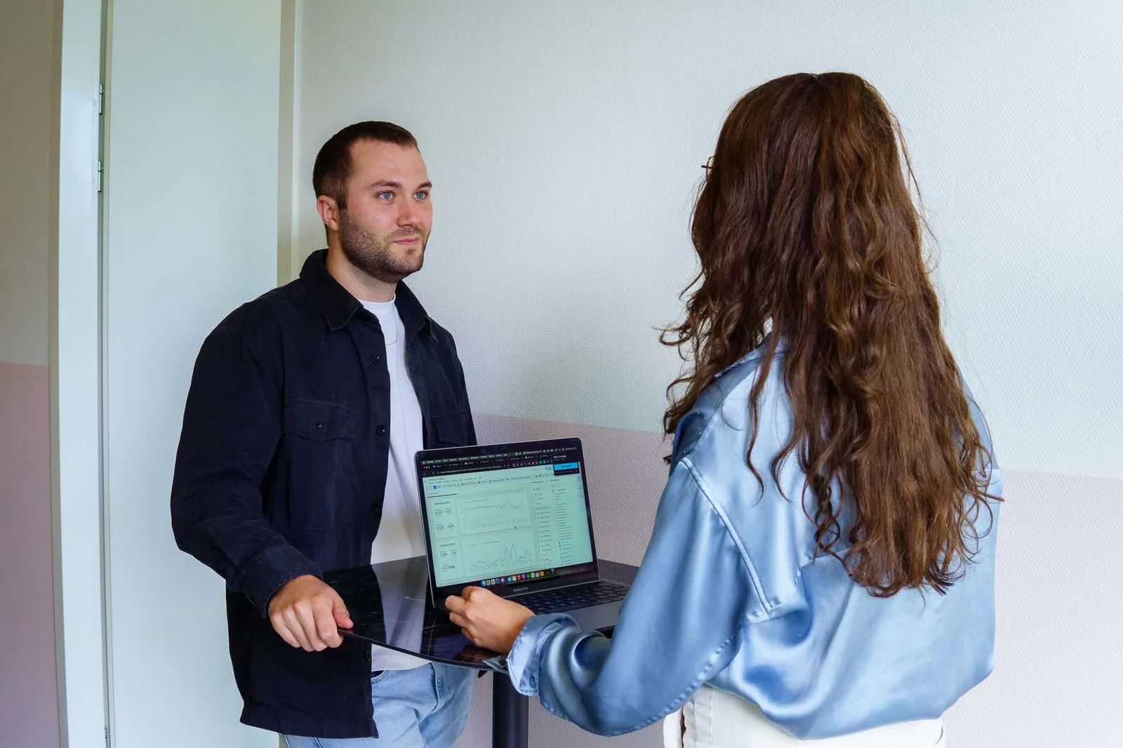 Twee jonge collega's bespreken data op een laptop in een lichte kantooromgeving, waarbij de vrouw de laptop vasthoudt en de man ernaar kijkt.