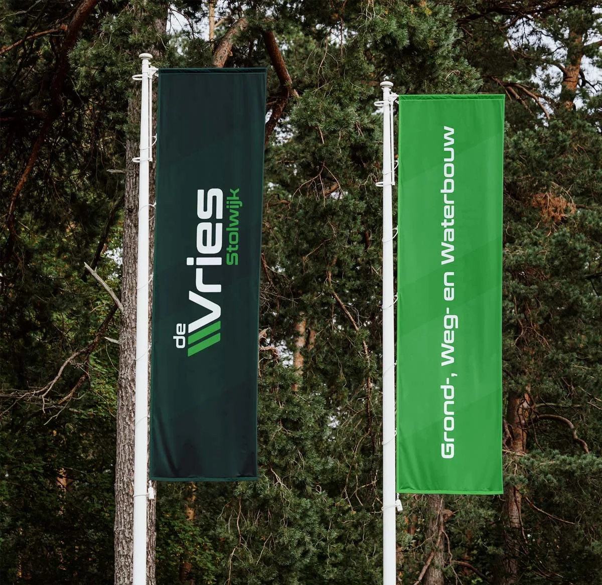 Twee vlaggen van De Vries Stolwijk, een donkergroene en een lichtgroene, wapperend voor bomen.